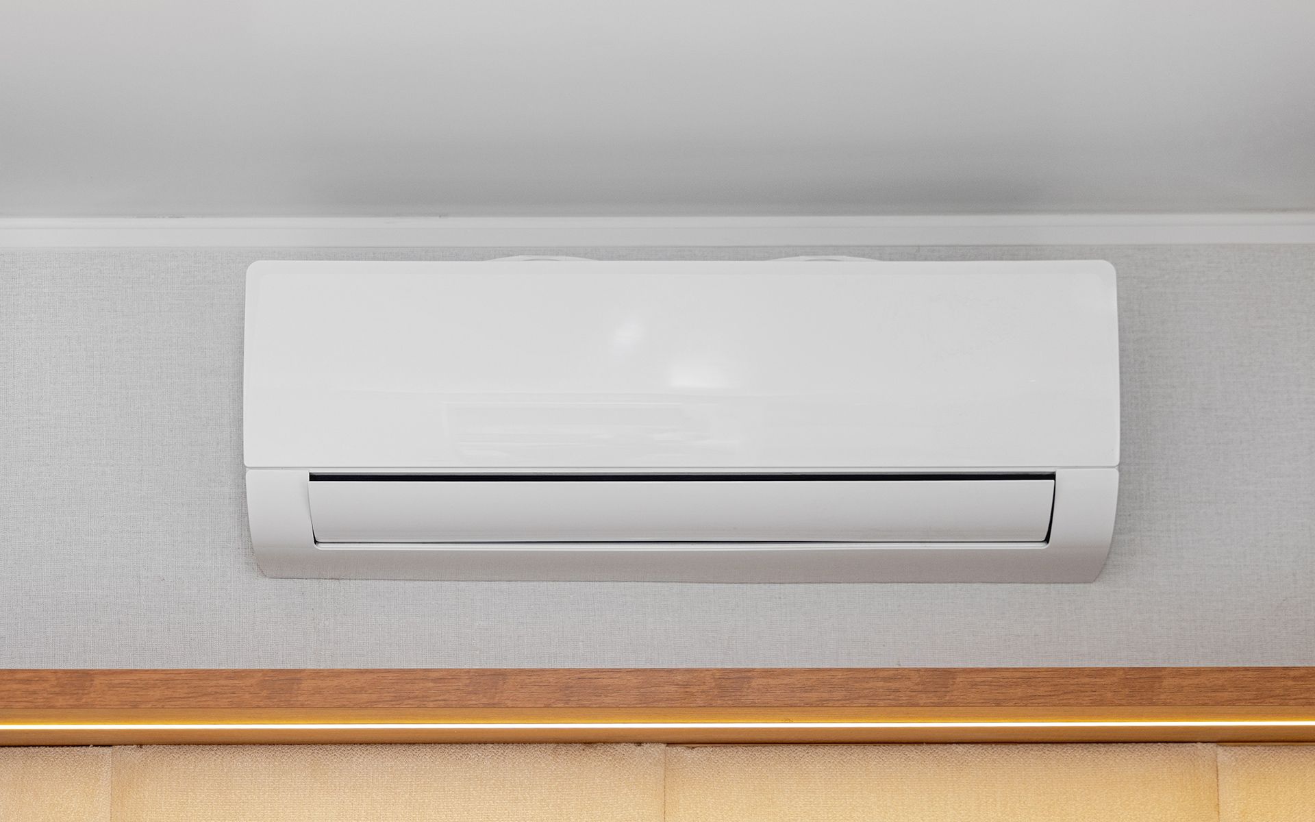 White wall-mounted air conditioning unit on a textured wall, above a wooden panel and window covering.