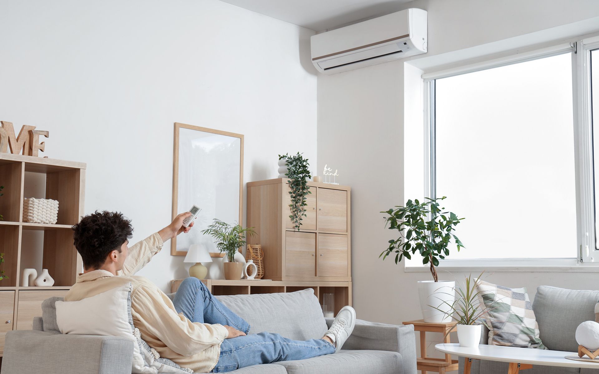 Person using air conditioner remote while relaxing on a sofa in a bright living room.
