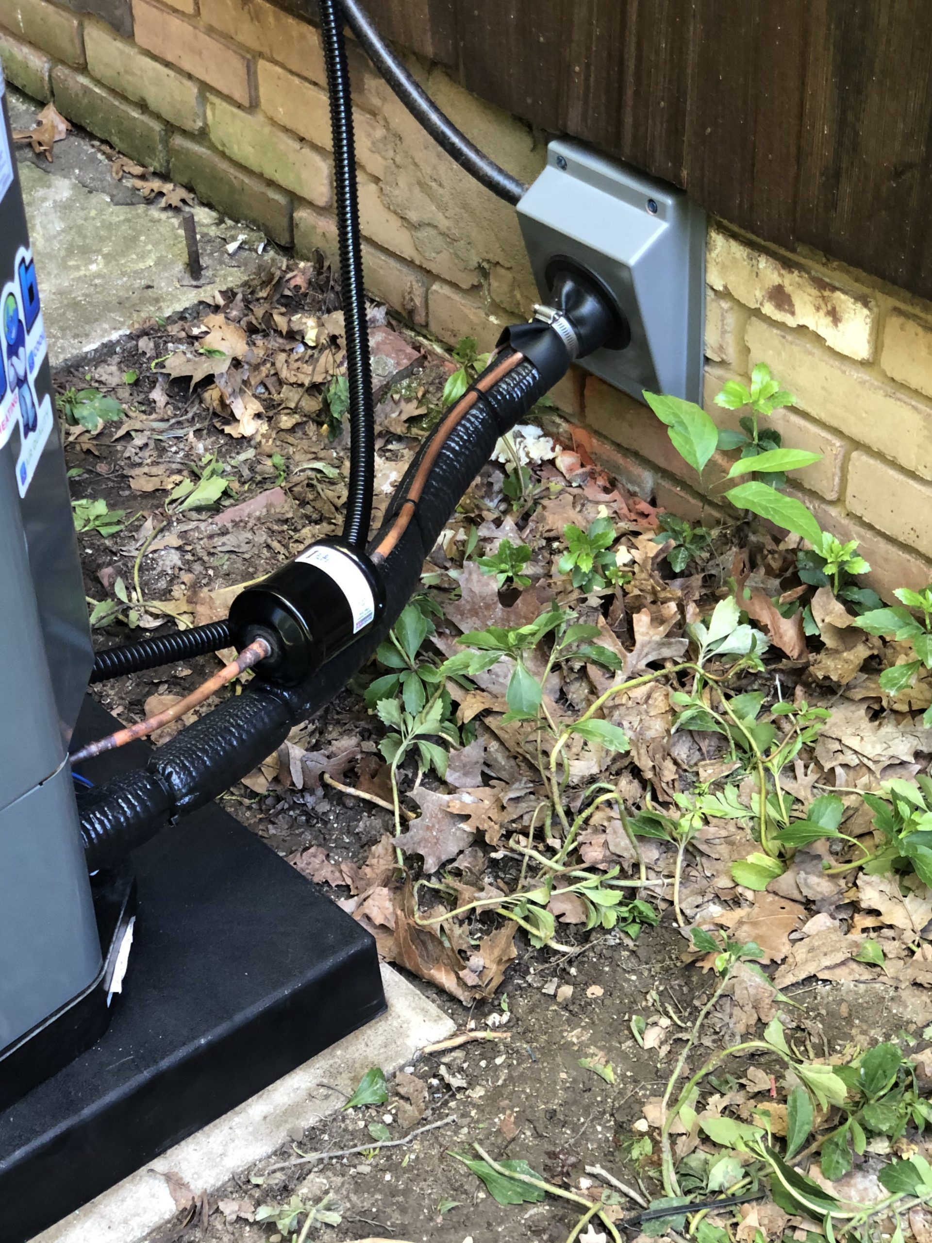HVAC unit next to a brick wall. Black and copper pipes are connected. Power outlet box is visible.