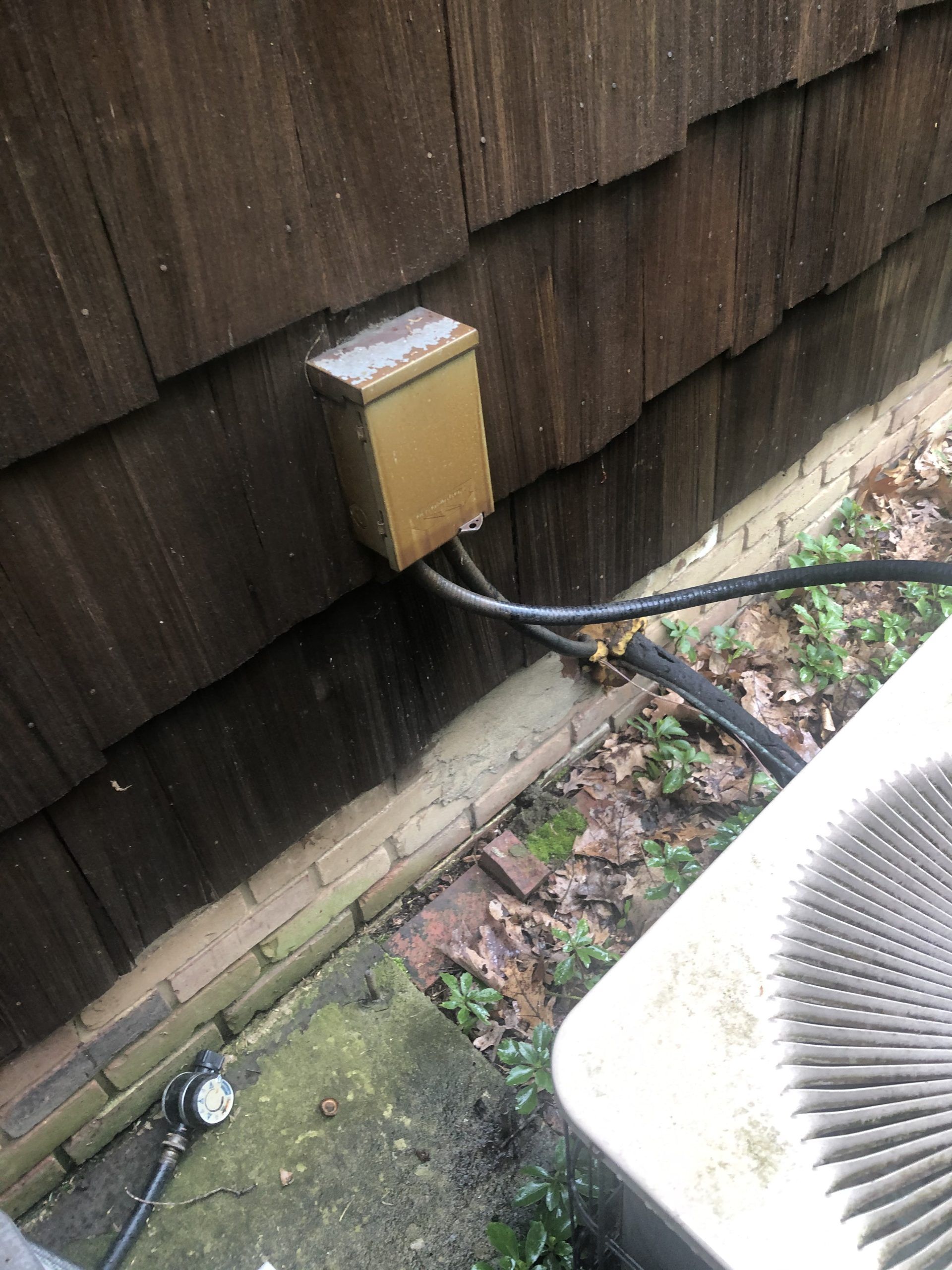 A brown electrical box is mounted on wood siding near an air conditioning unit.