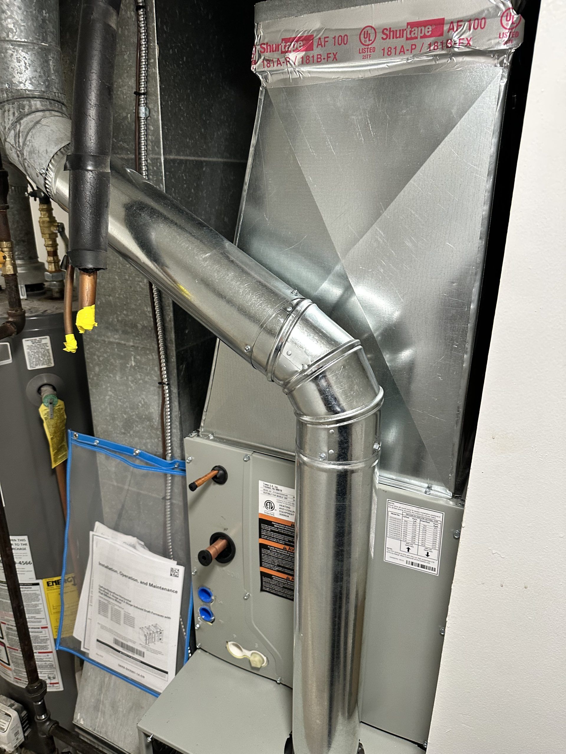Furnace and ductwork installation in a utility closet with metal and insulated pipes.