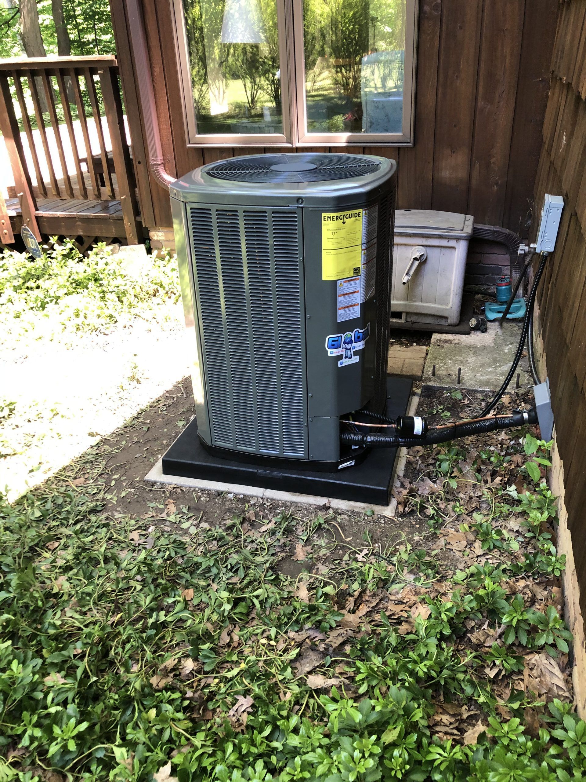 Outdoor air conditioning unit on a black platform, next to a building and overgrown greenery.