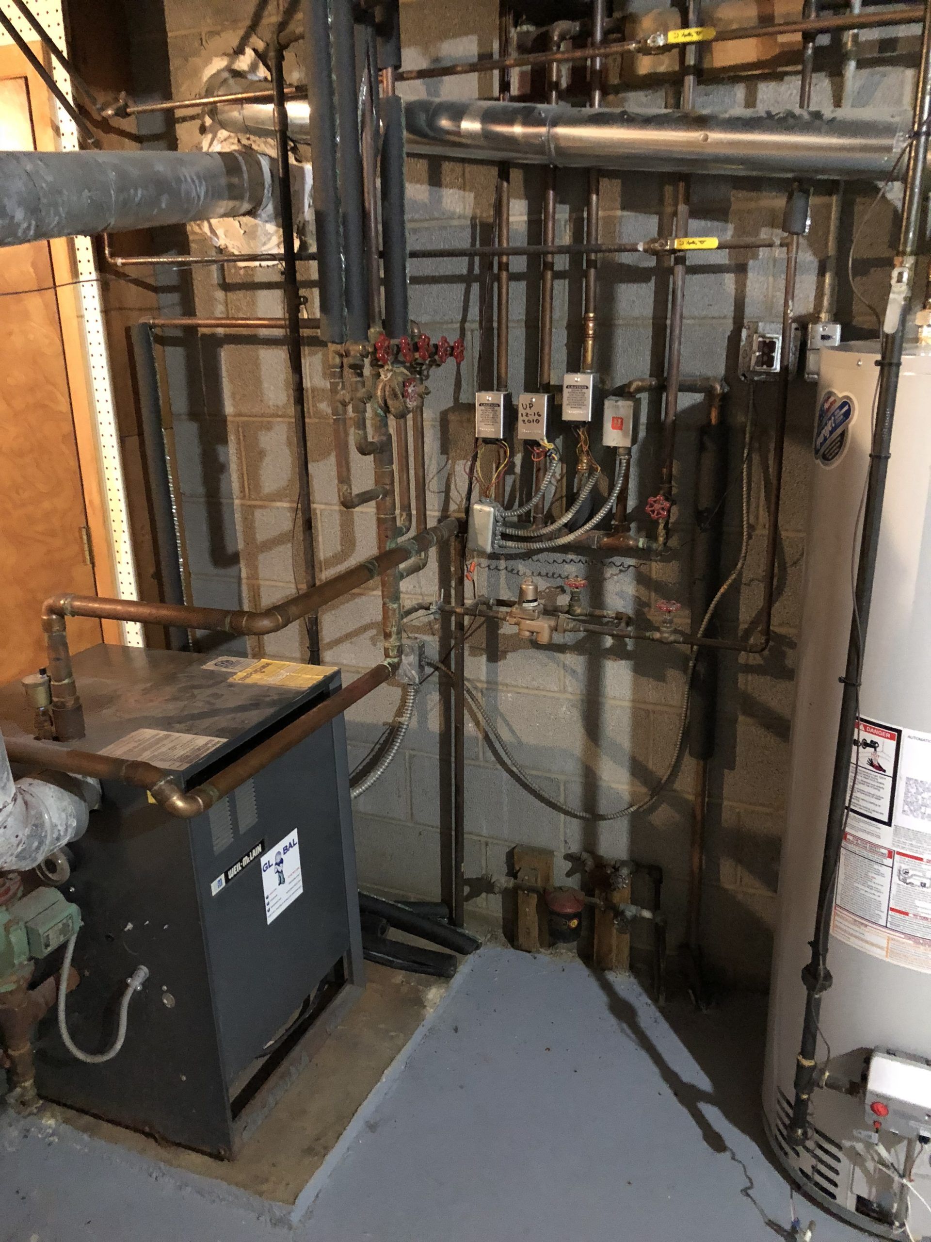 Basement mechanical room with pipes, water heater, boiler, and controls against a concrete block wall.