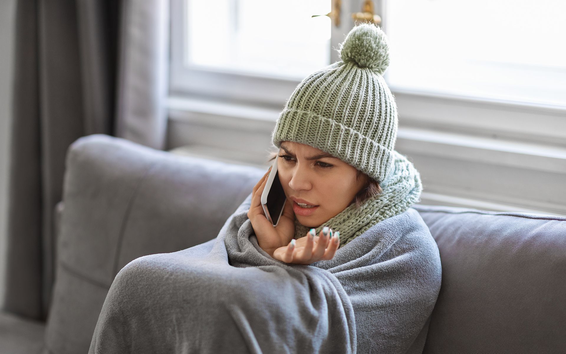 Woman wrapped in a blanket, wearing a hat, talking on a phone, looking concerned while sitting on a couch.