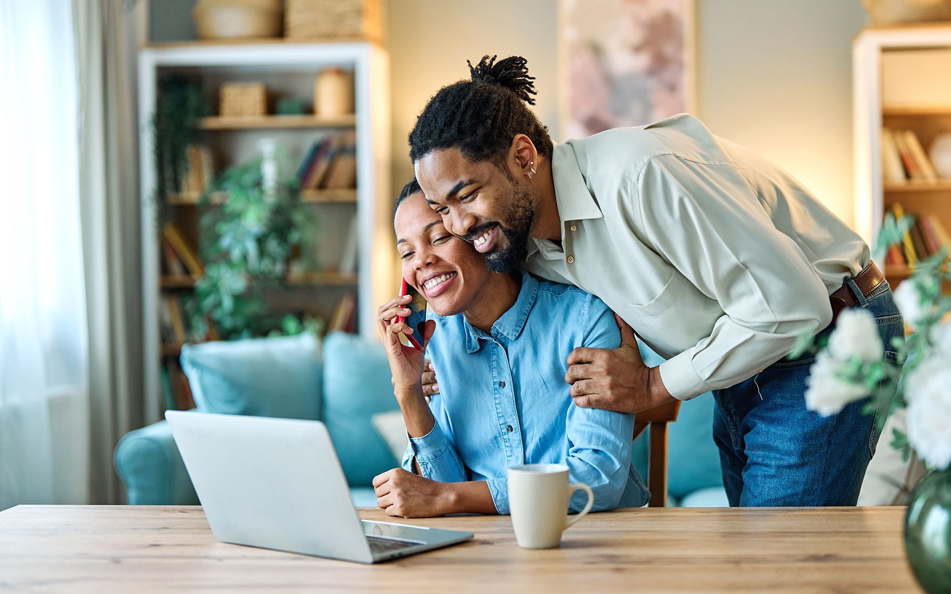 Couple working together, woman on phone and laptop, man hugging her, smiling in home.