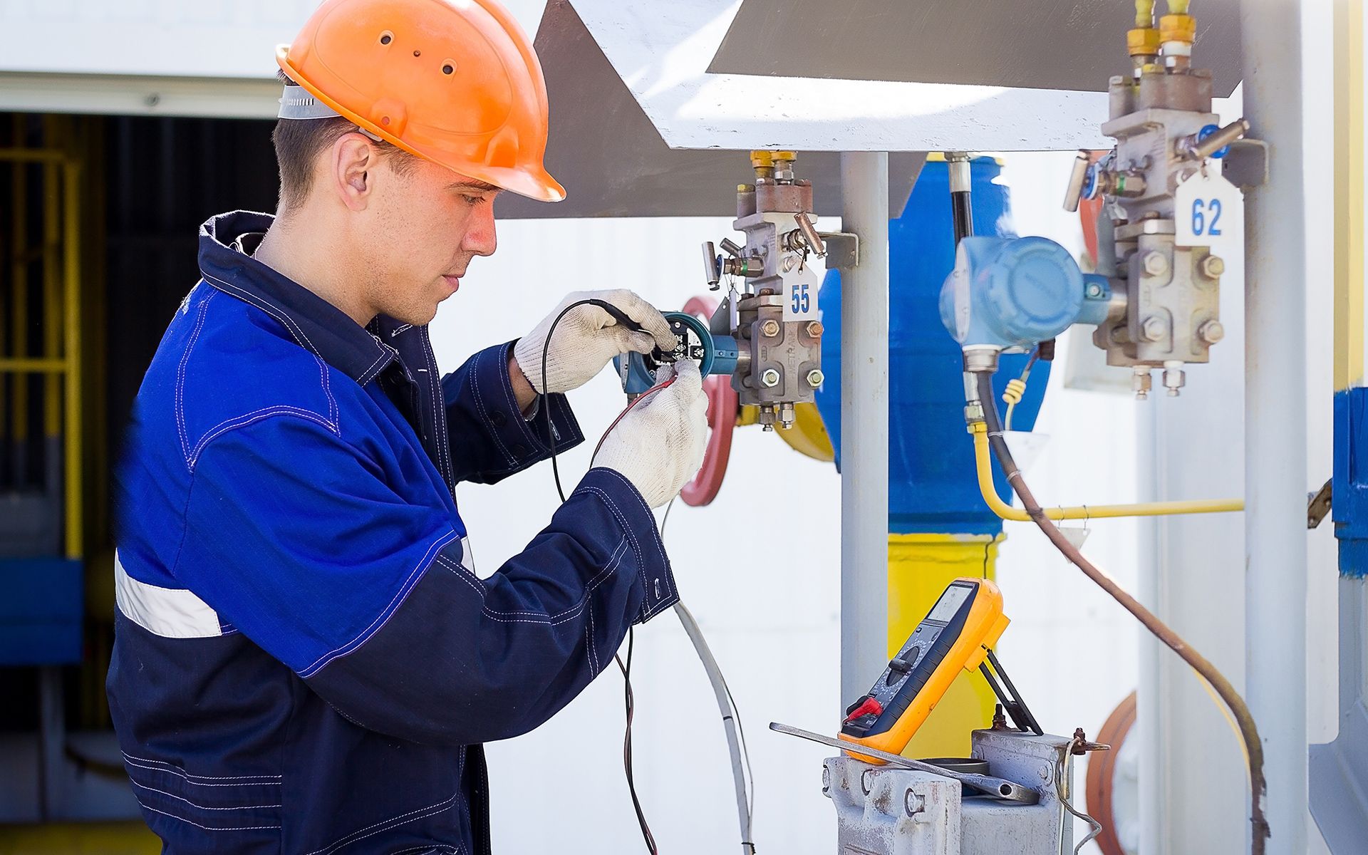 Technician in orange hard hat, blue jacket, and gloves, testing equipment with a multimeter.
