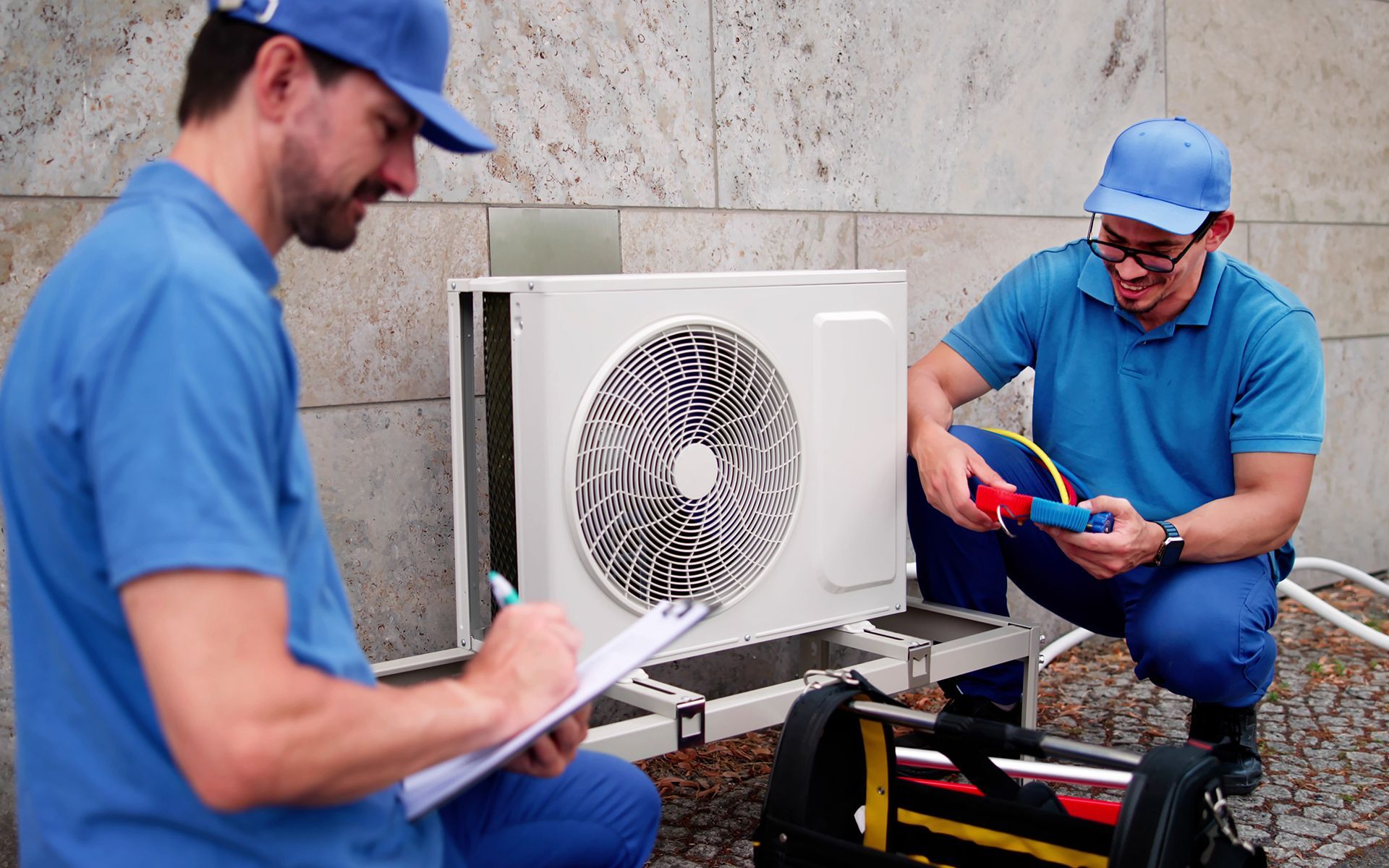 Two technicians in blue uniforms examine an AC unit outside. One writes on a clipboard, the other uses a meter.