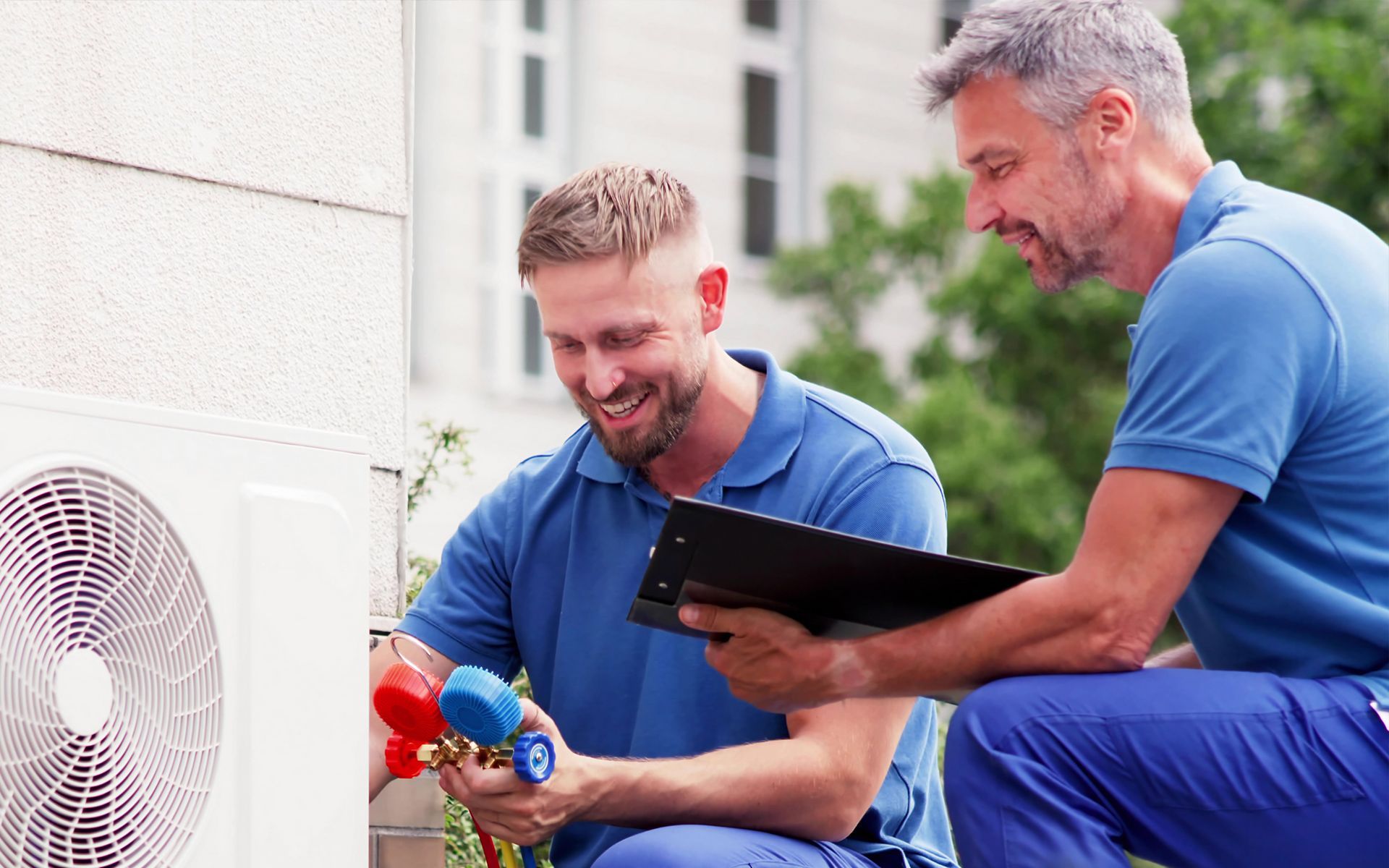 Two HVAC technicians working on an outdoor air conditioning unit. One holds gauges, the other has a clipboard, both smiling.