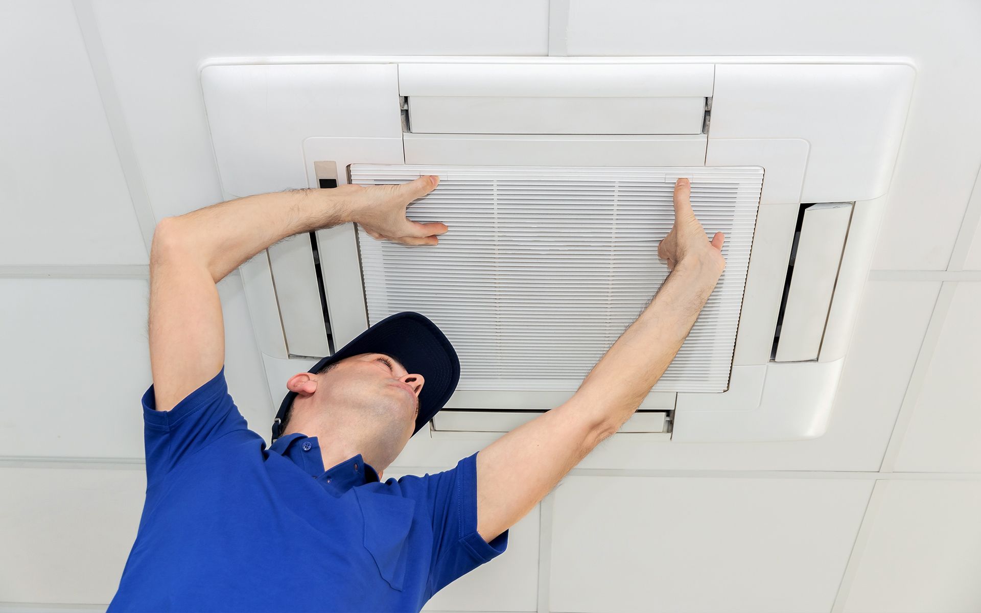 Man in blue shirt, black hat inspecting ceiling air vent, pointing to it.