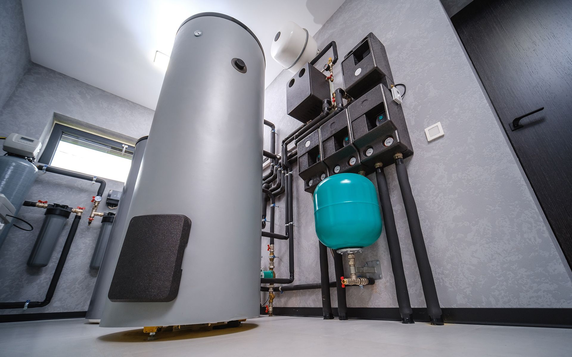 A water heater and associated plumbing in a utility room. Gray tank, blue tank, black pipes, light walls.
