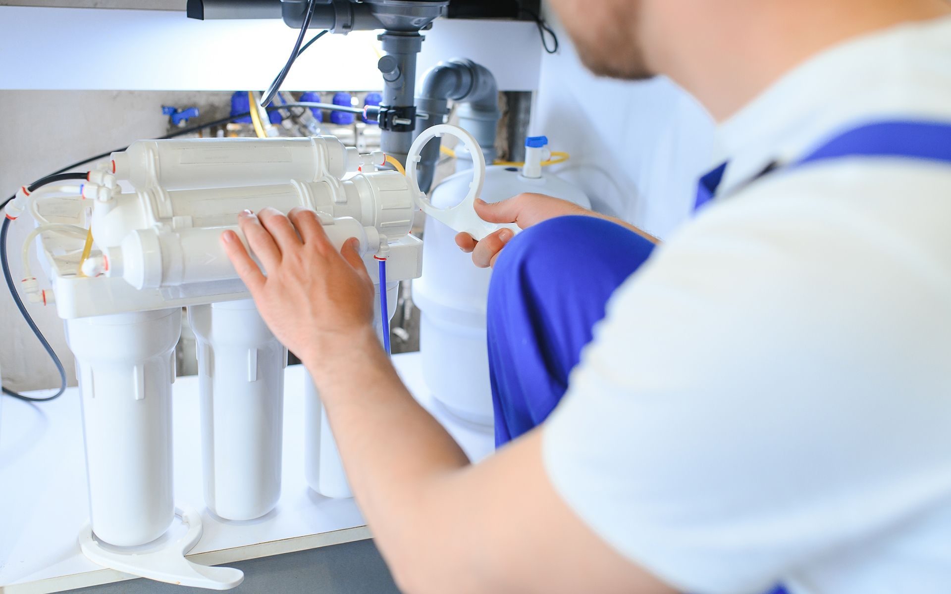Plumber working on a water filtration system under a sink, wearing blue overalls.