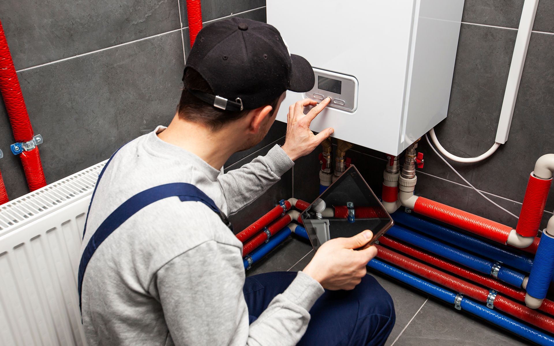 Plumber inspecting a white boiler, using a tablet, with red and blue pipes in a gray room.