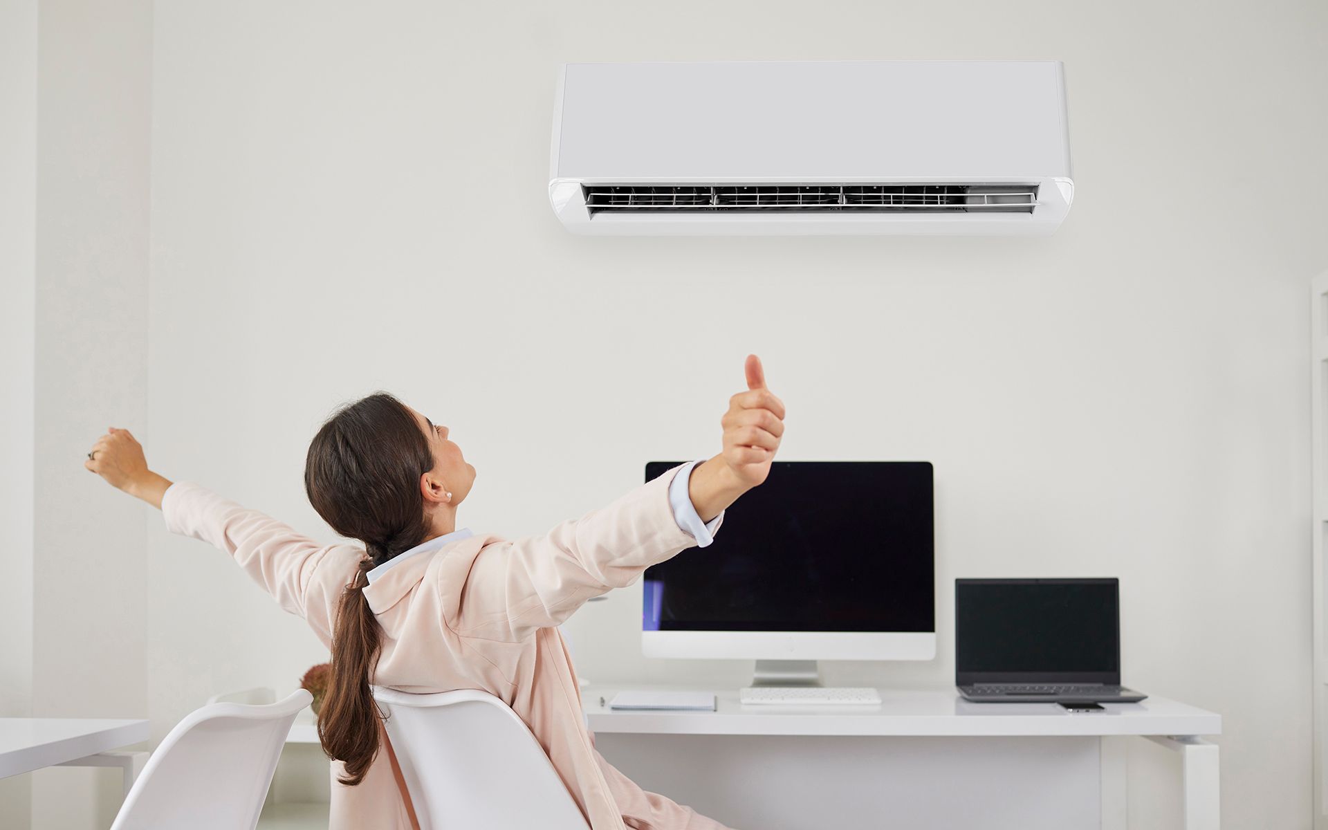 Woman celebrating under an air conditioner in an office, thumbs up, arms raised, by a desk.
