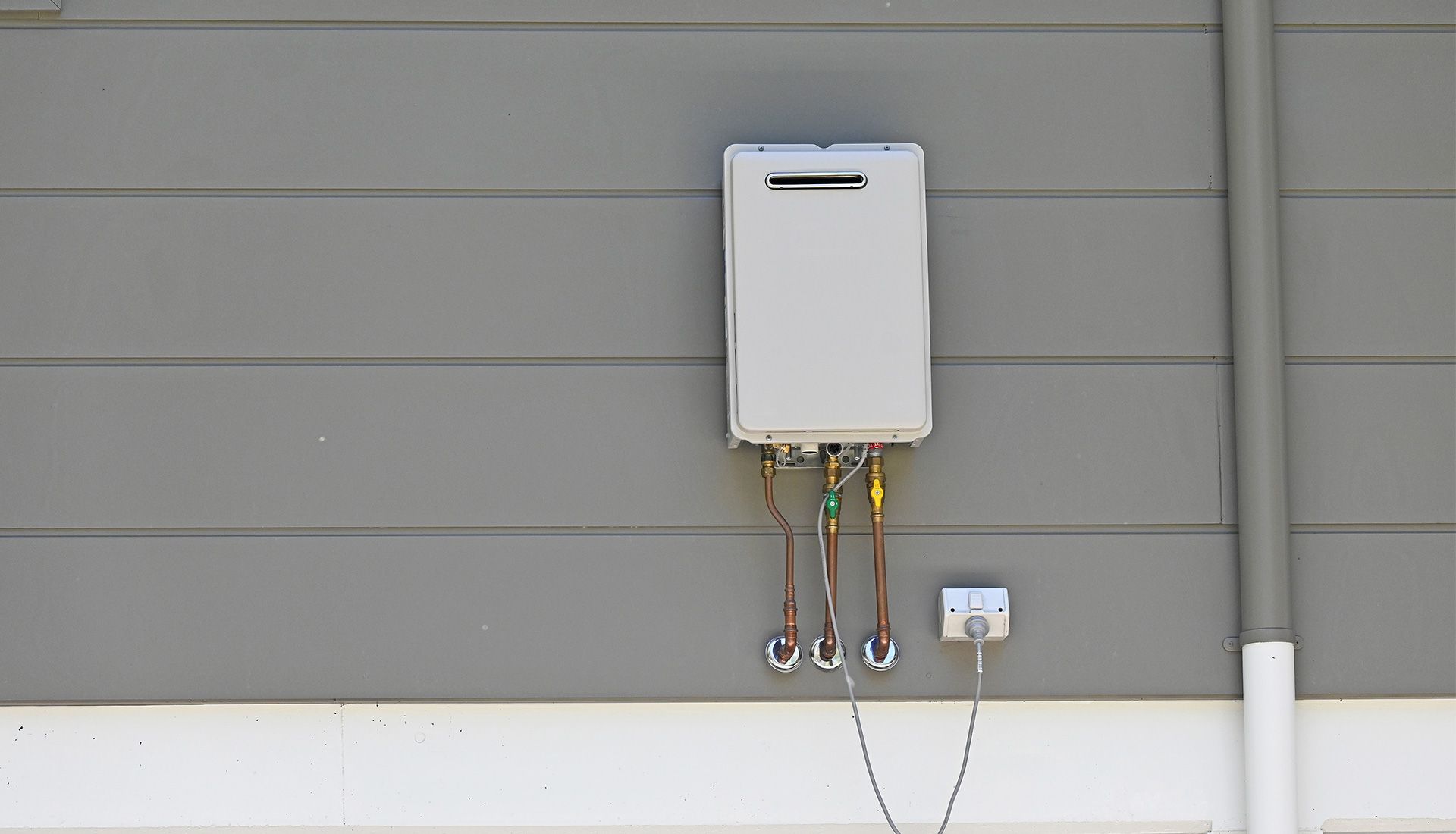 A grey, rectangular tankless water heater mounted on a grey house siding with copper pipes and electrical box.
