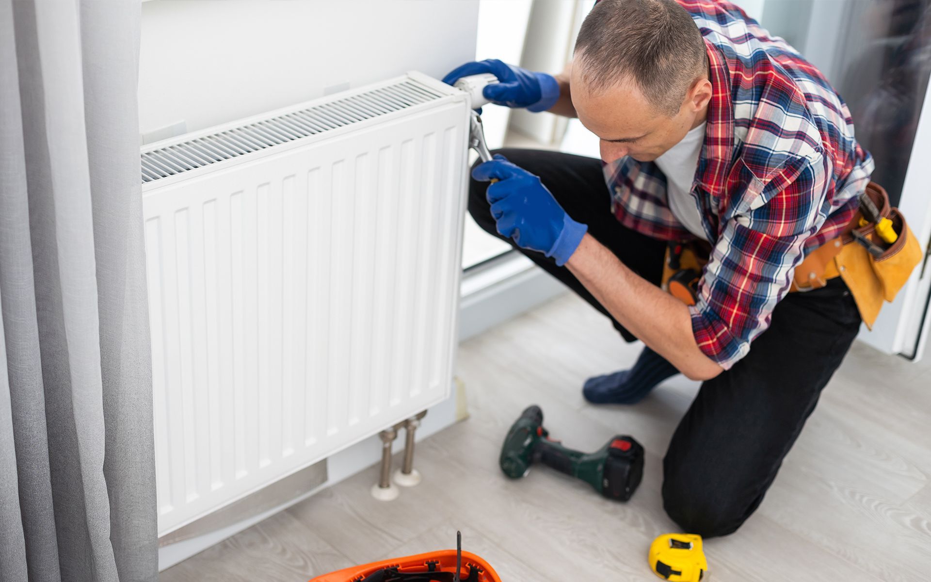 Man in blue gloves works on a white radiator. Drill and tools nearby.