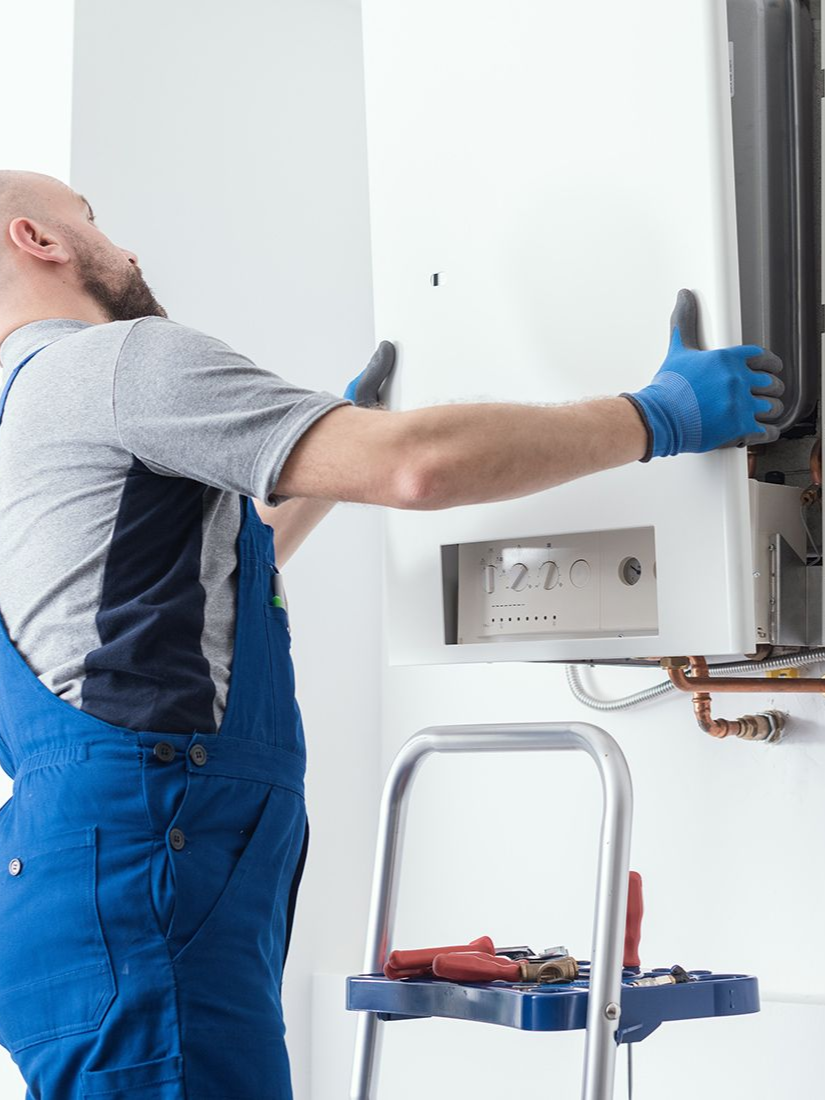 A person in blue overalls installing a white wall-mounted boiler, using a step stool.