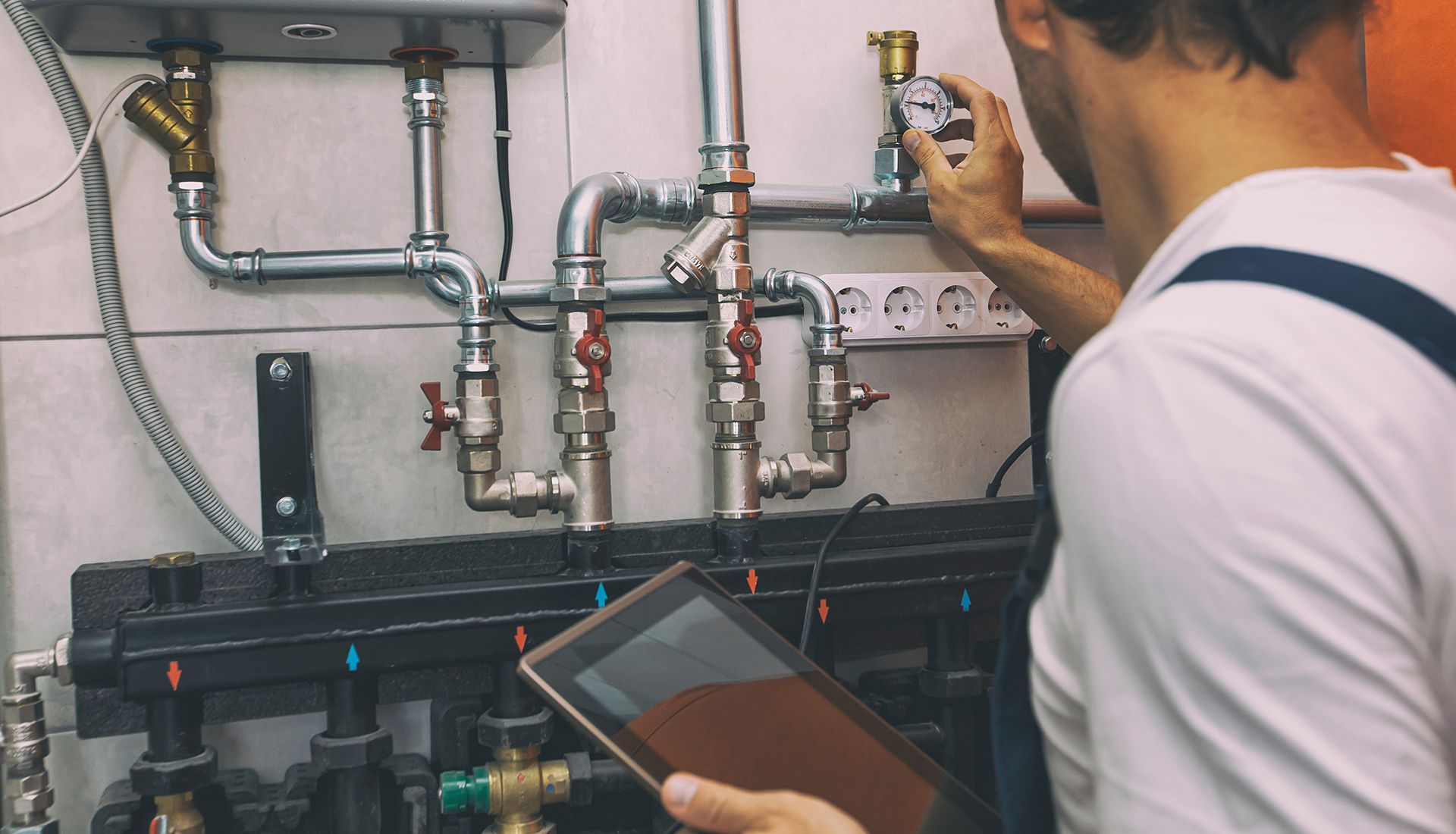 Plumber adjusting valves on pipes, using a tablet for diagnostics in a utility room.