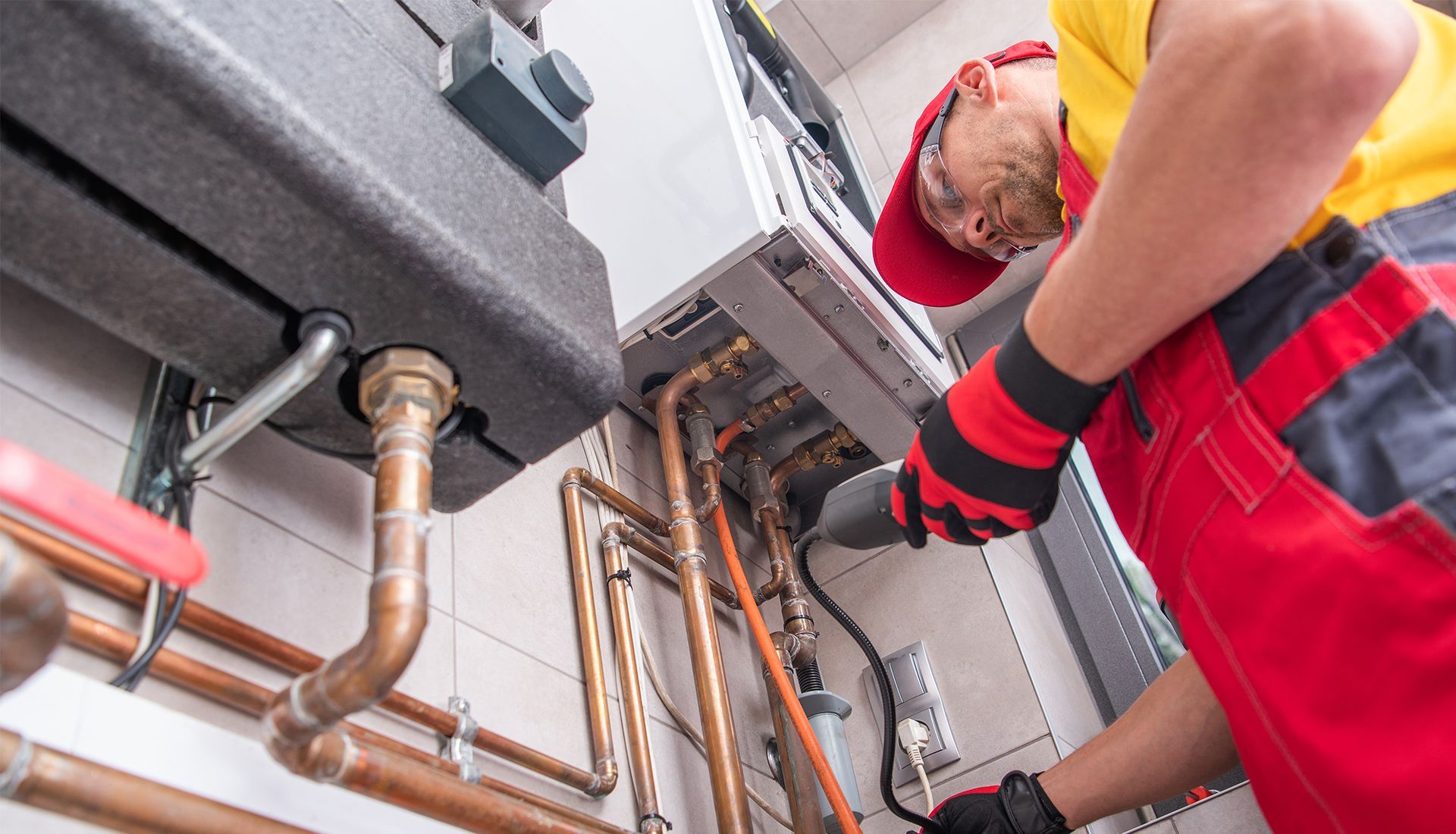 Plumber in red overalls inspecting a boiler. Copper pipes and machinery visible.