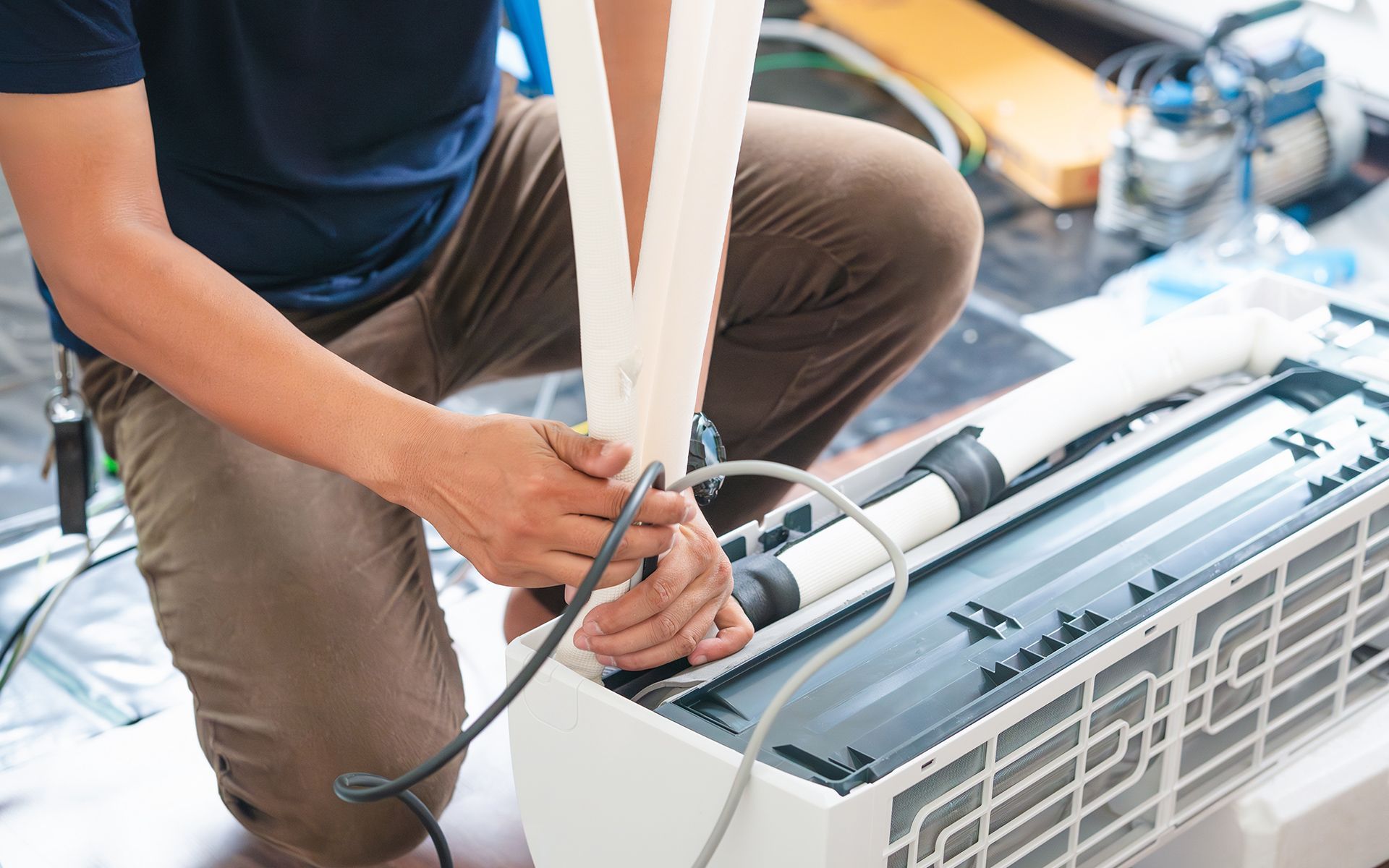 Person kneeling, installing air conditioner unit, attaching hoses, in a room.