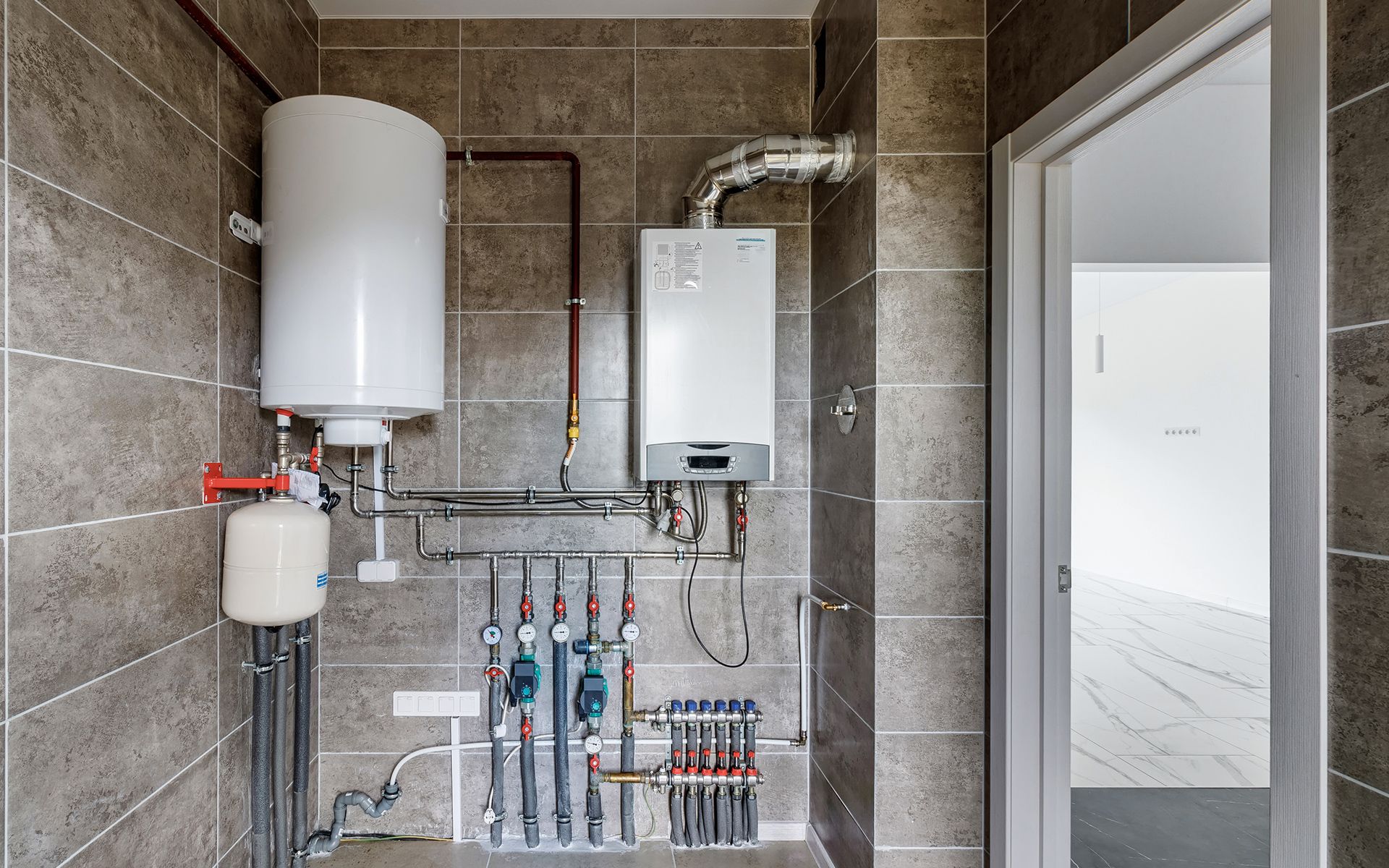 Water heater and furnace mounted on a tiled wall in a utility room, near an open doorway.