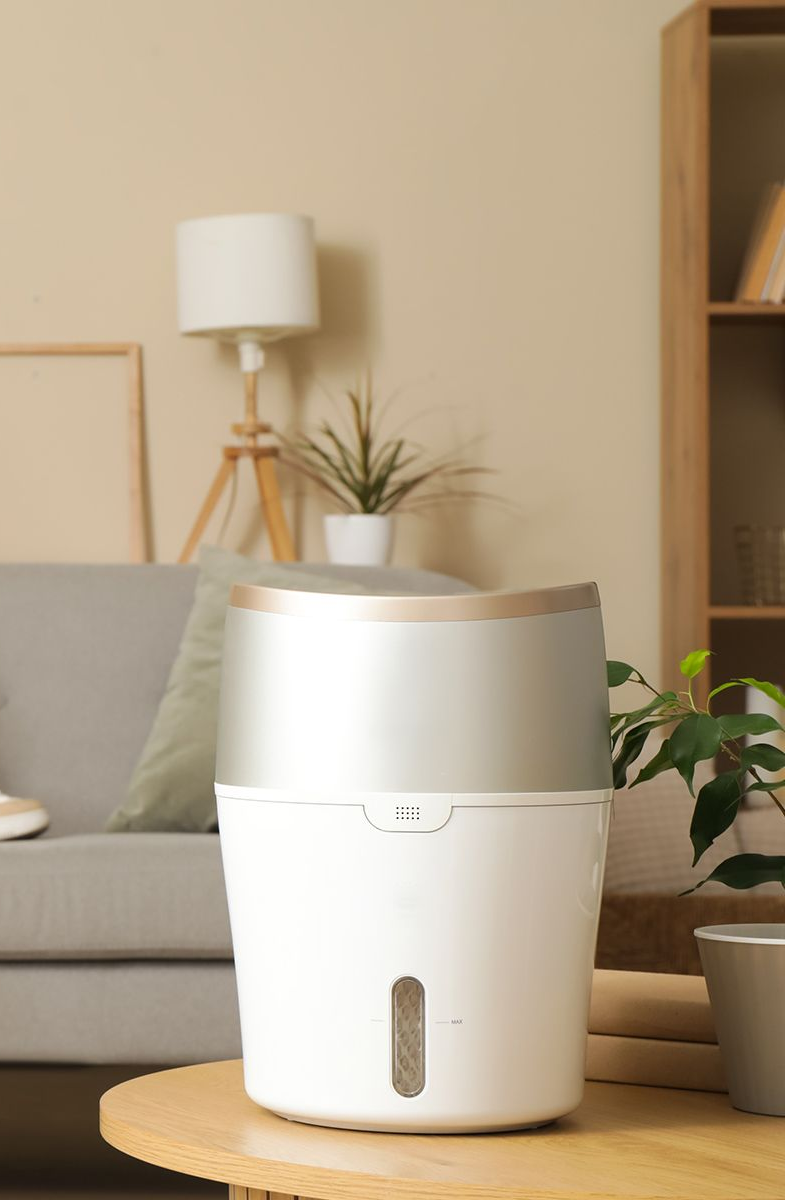 White and gold humidifier on a round wooden table in a living room, sofa and lamp in background.