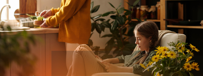 Woman relaxing while another person prepares food in a kitchen. Yellow flowers are in front.