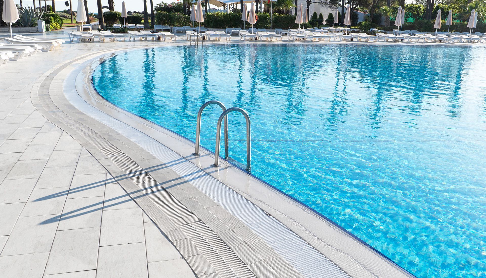 Curving blue pool with metal ladder, surrounded by white tiles and lounge chairs. Sunny outdoor setting.