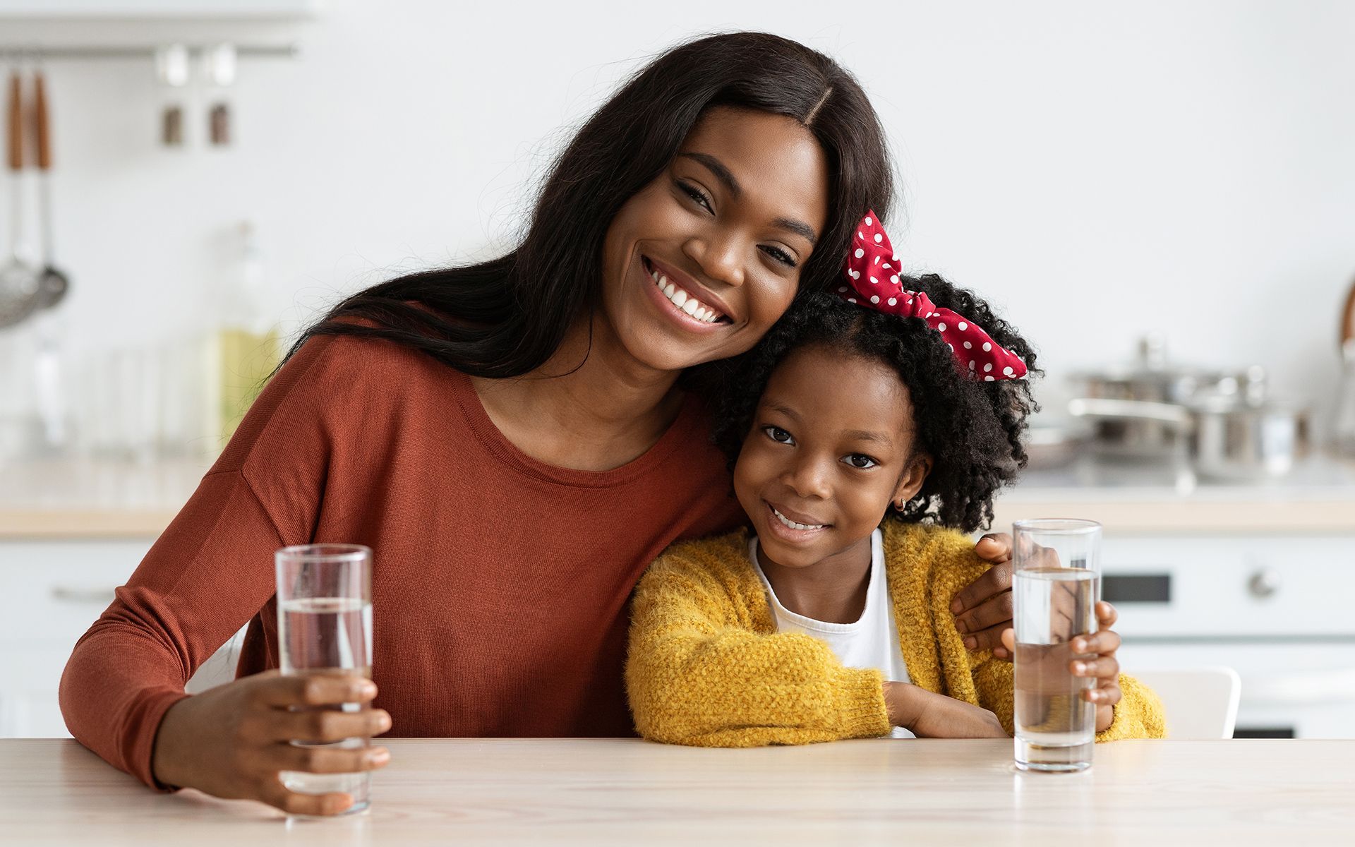Woman and child smiling, holding glasses of water, kitchen setting.