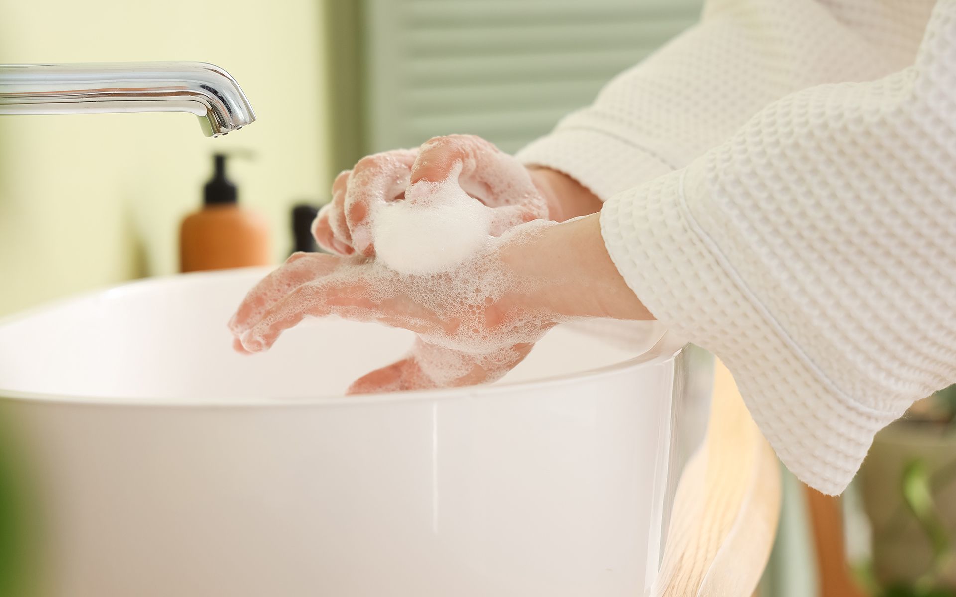 Hands washing with soap at a white sink, faucet on. Person wearing a white robe.