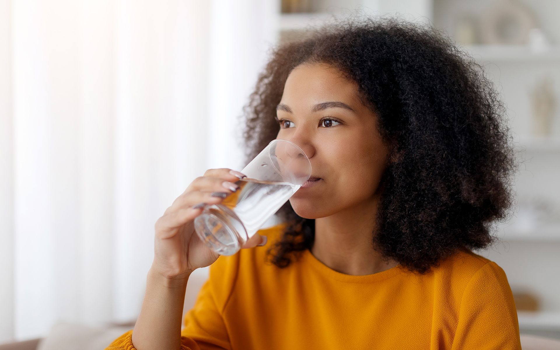 Woman drinks water from a clear glass, looking to the side with curly dark hair and wearing a yellow shirt.