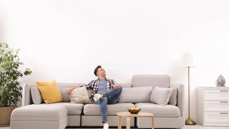 Man relaxing on a grey sectional sofa, looking upwards in a bright living room.