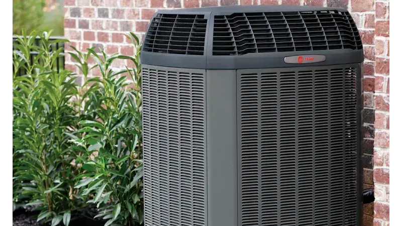 Dark grey air conditioning unit next to a brick wall and green shrubs.