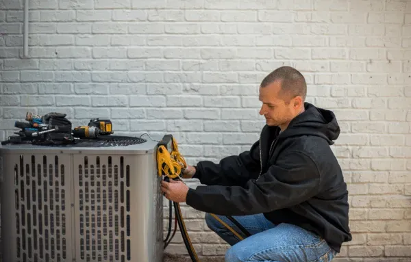 Man inspects a white heat pump unit installed outdoors next to a brick wall.
