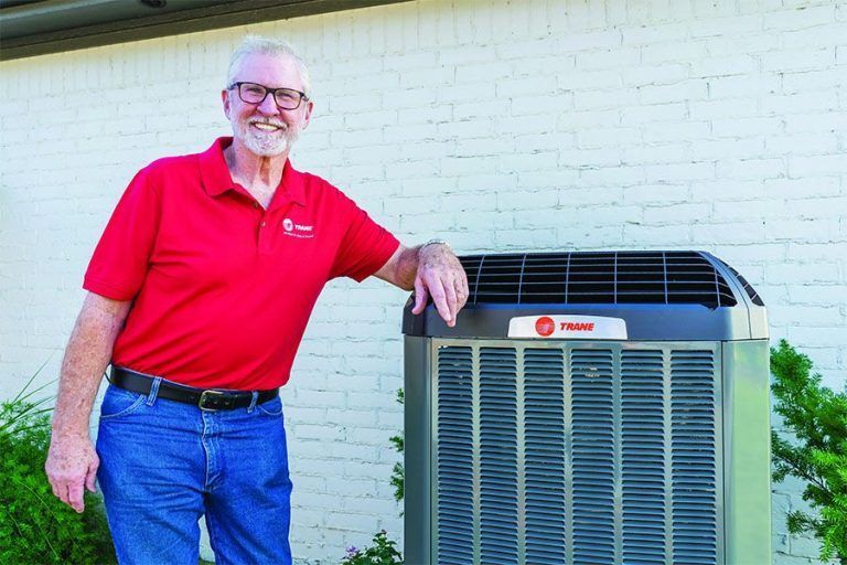 Man in red shirt and jeans leaning on an air conditioning unit outside a building.
