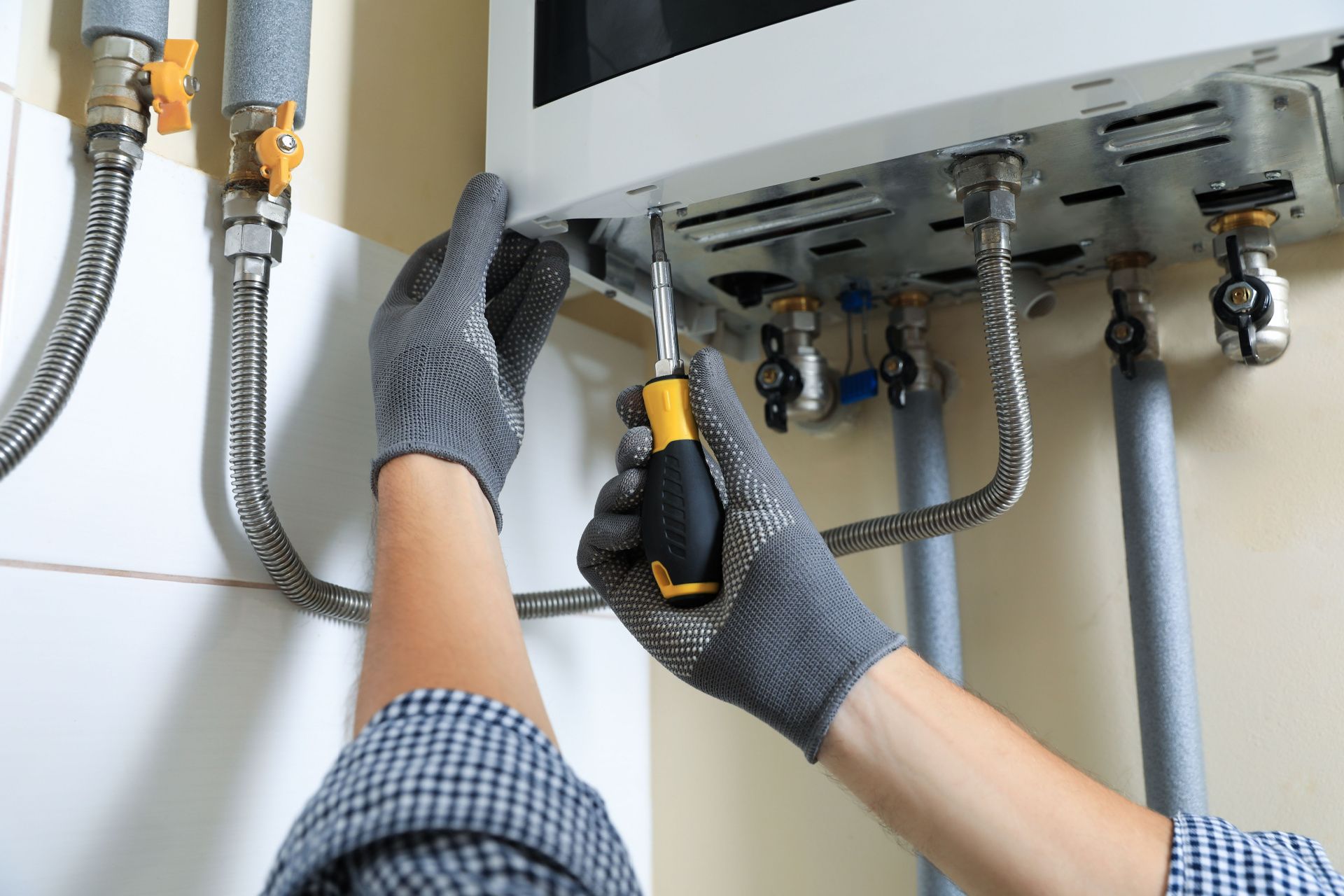 Man in a cap examines a white wall-mounted boiler, possibly adjusting settings while holding a tablet in a utility room.