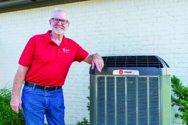Man in red shirt and jeans leaning on Trane air conditioning unit outside a white brick building.