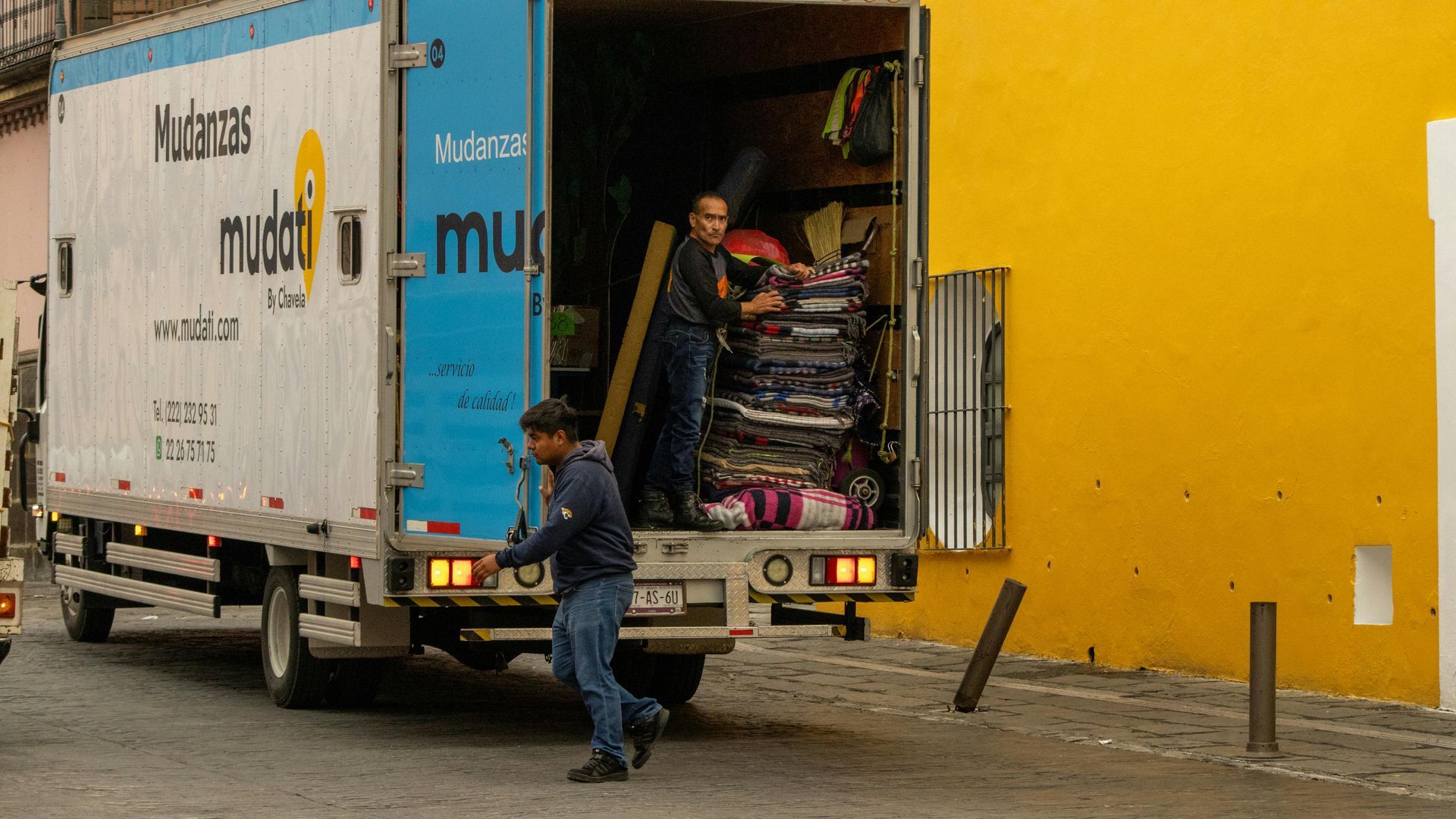 A man is loading a moving truck with a yellow building in the background.