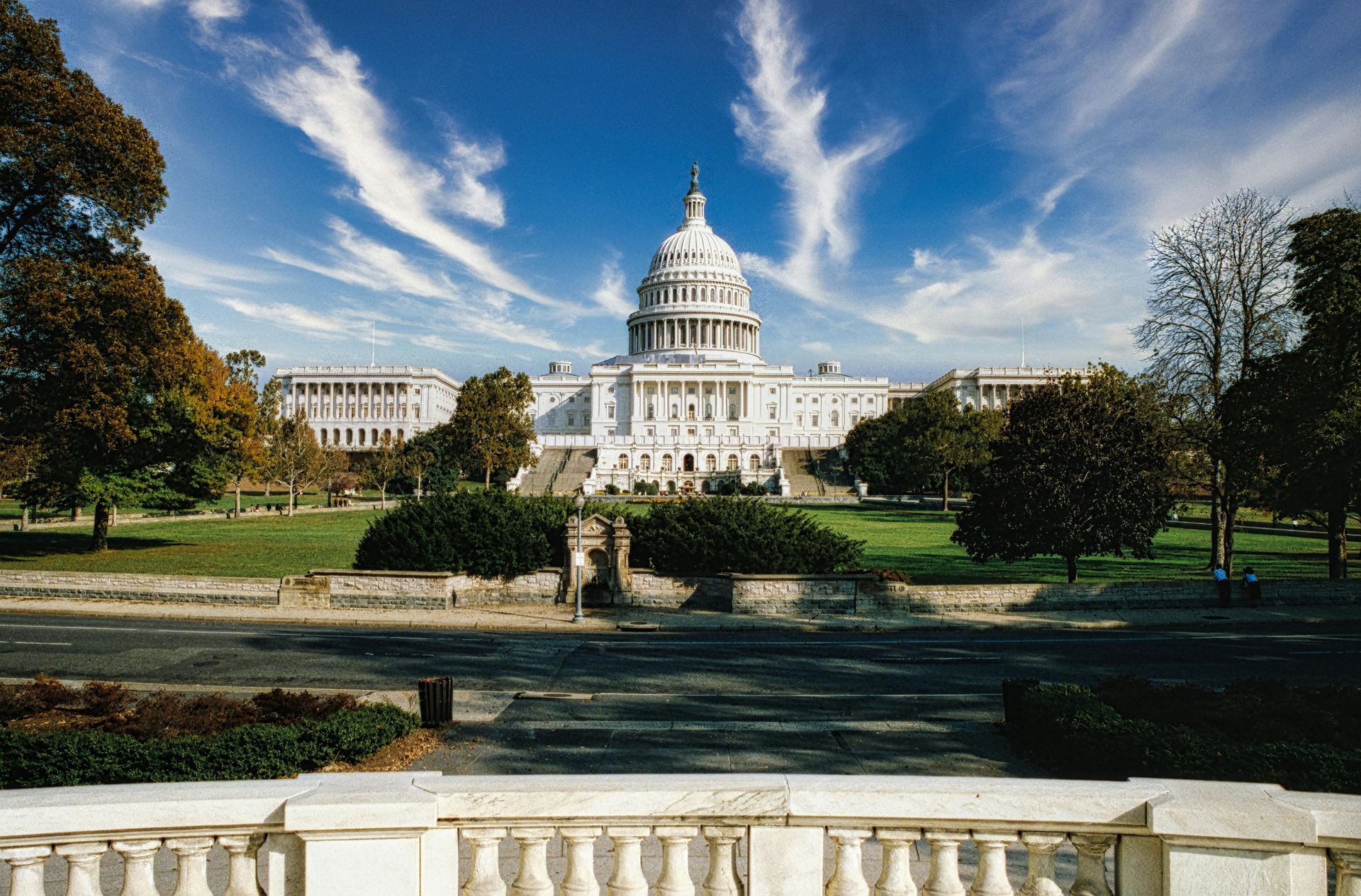 U.S. Capitol building under a blue sky, viewed from a formal garden with trees and a reflecting pool