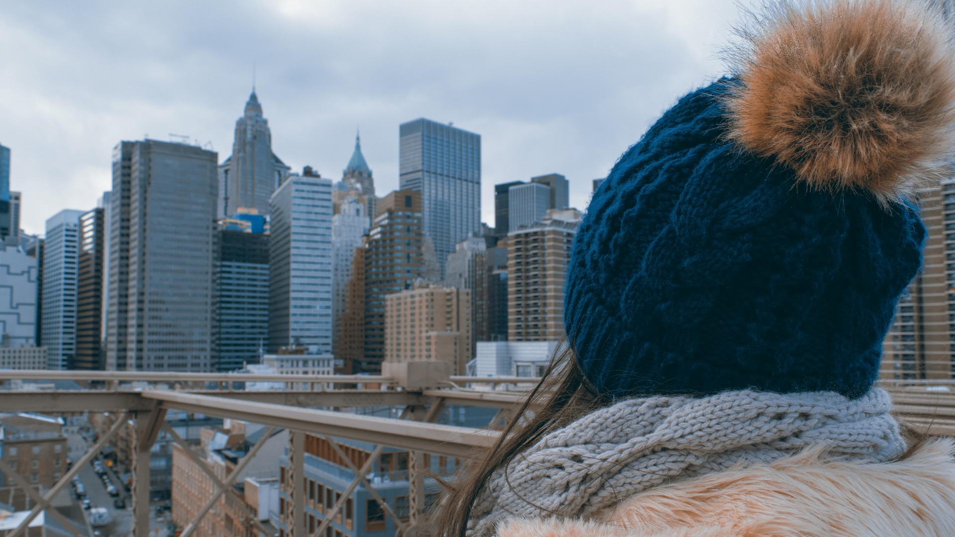 Person in blue knit hat and scarf overlooking a snowy city skyline from a bridge.