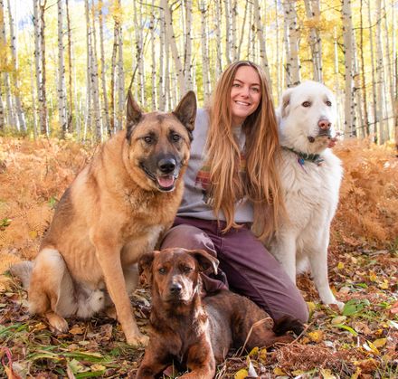 A woman is kneeling down with two dogs in the woods