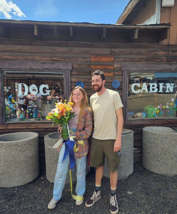 A man and a woman are standing in front of a dog cabin holding flowers.