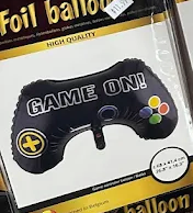 Black game controller-shaped foil balloon with “GAME ON!” text, colored buttons, and yellow plus symbol in packaging