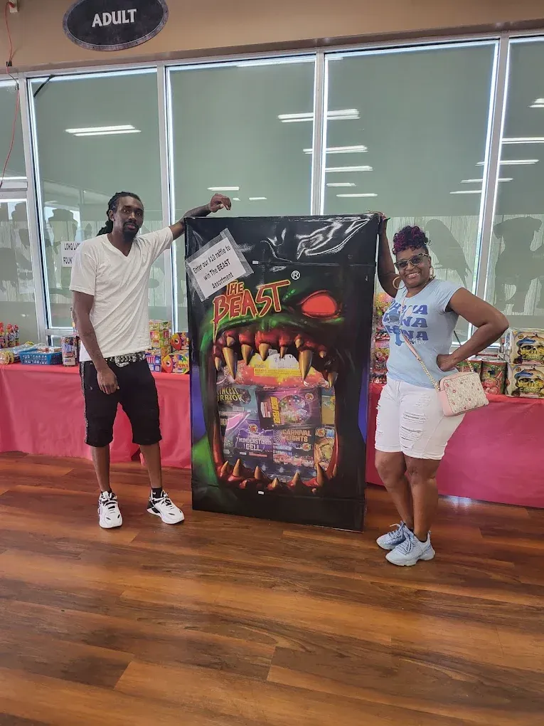 Two people posing beside a colorful arcade game display in a hall with wooden floors.