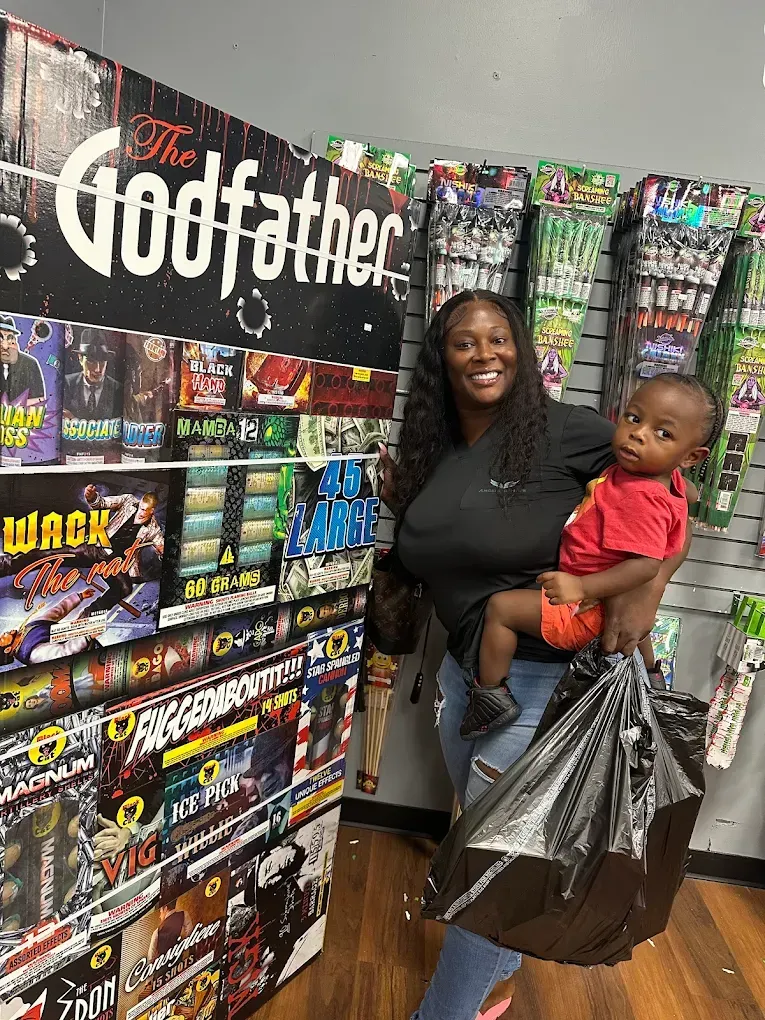 Woman smiling and holding a child in a store aisle with colorful fireworks displays and a Godfather sign