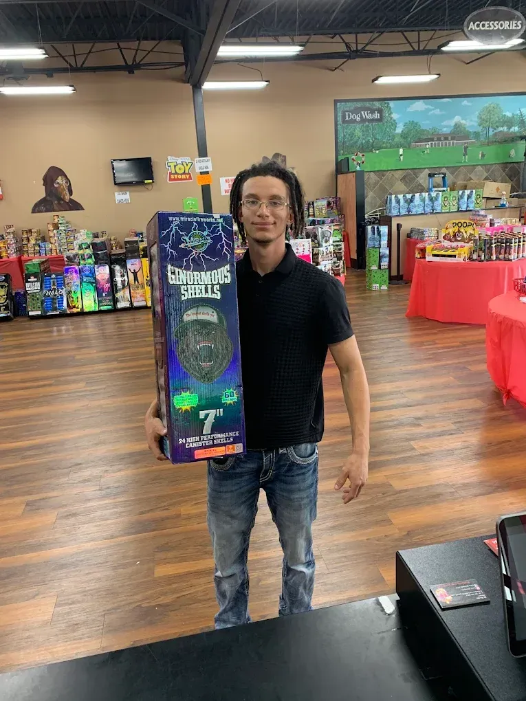 Man standing in a store aisle holding a tall colorful box, with toy displays and tables in the background.
