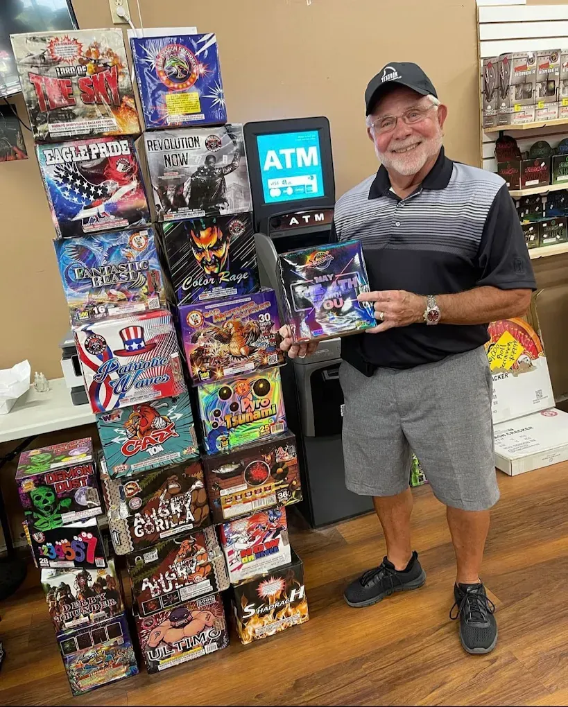 Man smiling beside a display of colorful trading card boxes in a store, holding a box and a sign labeled ATM.