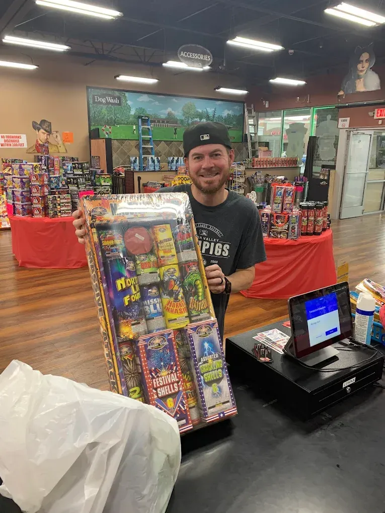 Smiling man at a fireworks store counter, holding a colorful display box beside a checkout register.