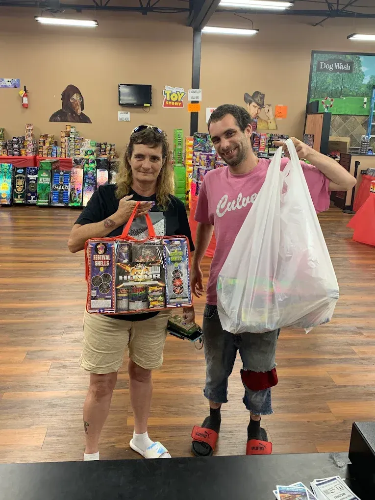 Two people pose in a store aisle holding large toy/game purchases and a shopping bag.