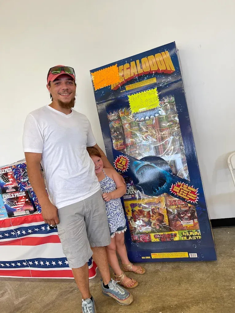 Man standing beside a colorful fireworks display in a bright room, smiling and wearing a white shirt and cap