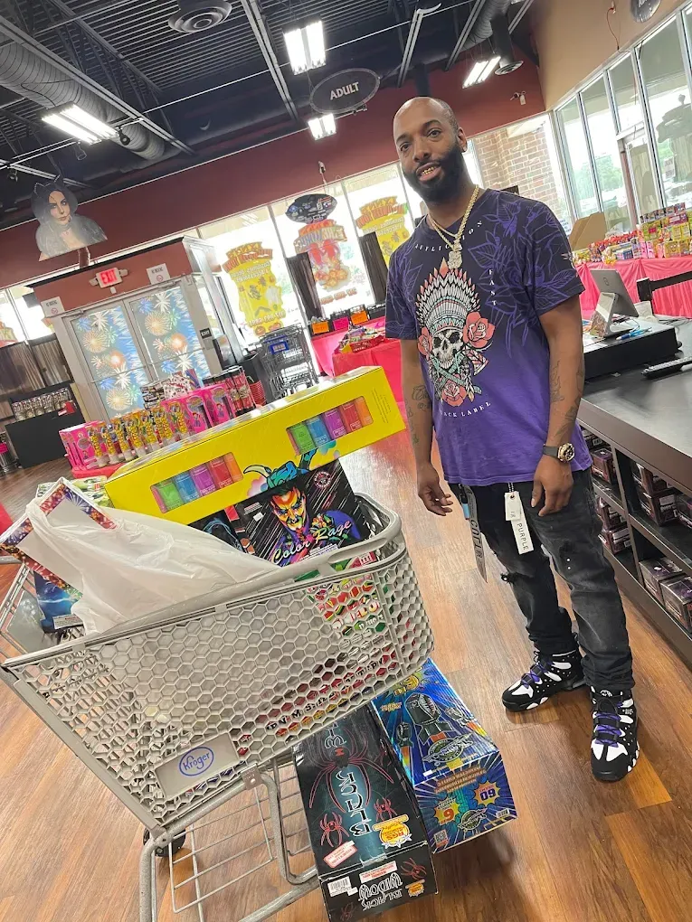 Man standing beside a shopping cart in a brightly lit store aisle, wearing a purple shirt and black pants.